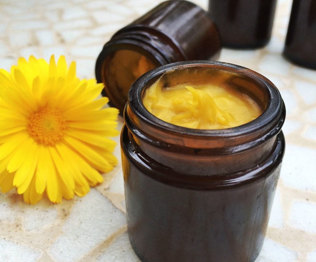 brown glass jar filled with thick yellow calendula salve with a yellow calendula flower next to it