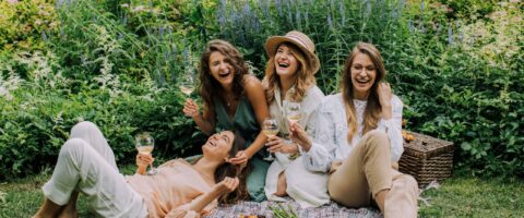 Four women enjoying a picnic, laughter, and wine in a vibrant summer garden.