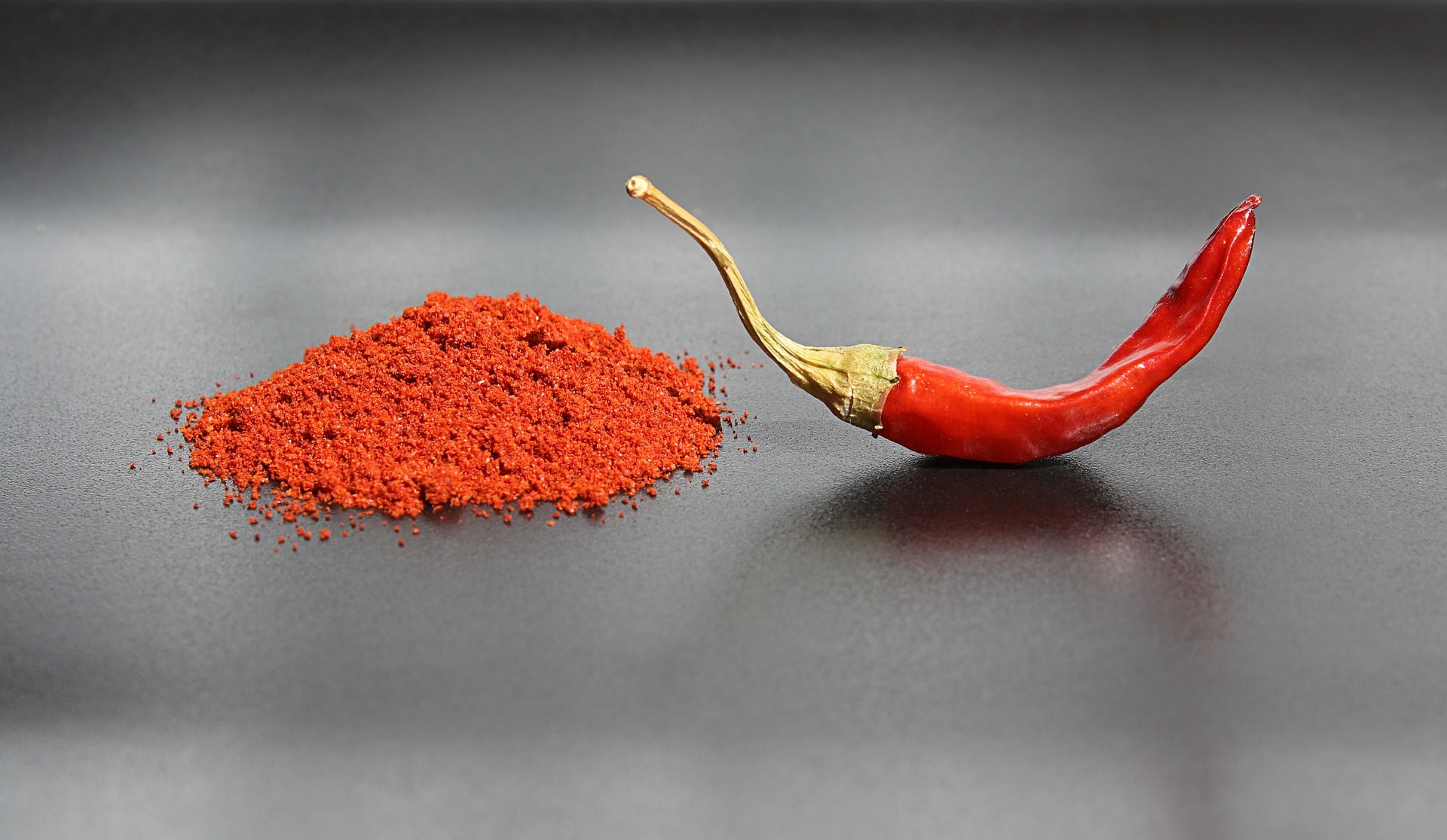 pile of red pepper powder on the left and a red chili pepper on the right on a gray background